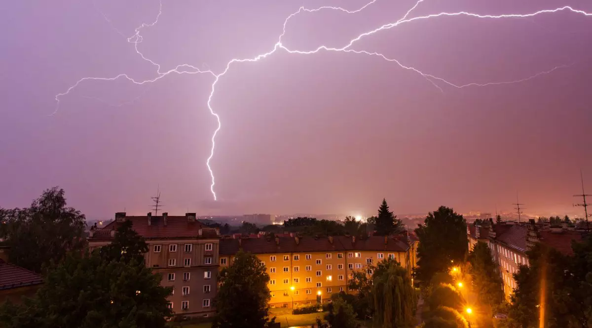 Furtună cu fulgere peste un oraș, pe timp de noapte, cu cerul aproape violet din cauza fulgerelor puternice care lovesc pământul