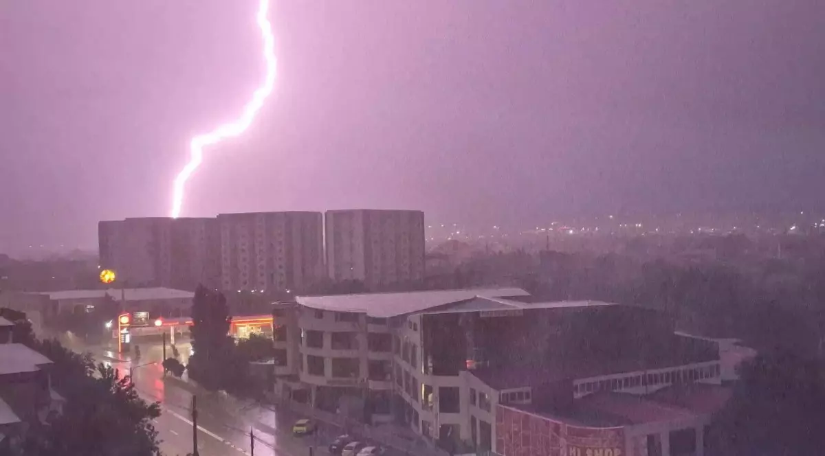 Imaginea surprinde un fulger puternic care luminează cerul violet al nopții, lovind în apropierea unor blocuri de locuințe dintr-o zonă urbană pe timp de furtună