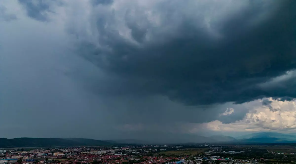 Cer plin de nori negri e furtună, deasupra unui oraș și a unei zone de relief muntoase în depărtare.