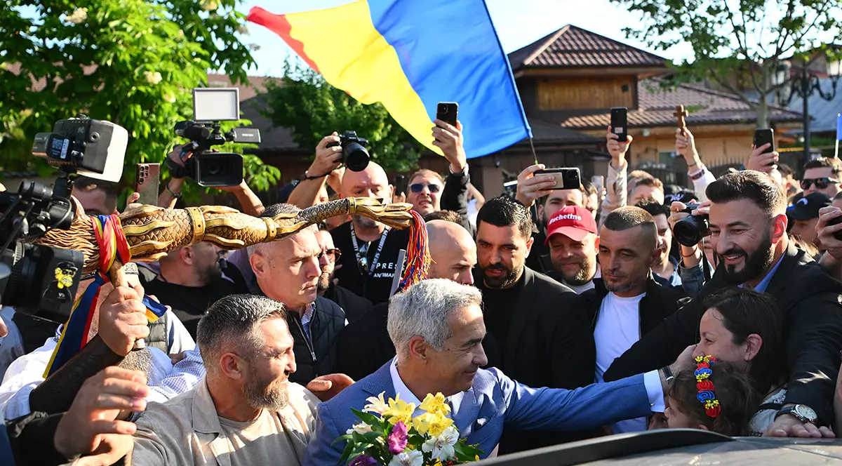 Călin Georgescu îmbrăcat într-un costum albastru deschis cu flori în mână, iar în spate susținătorii flutură un steag.