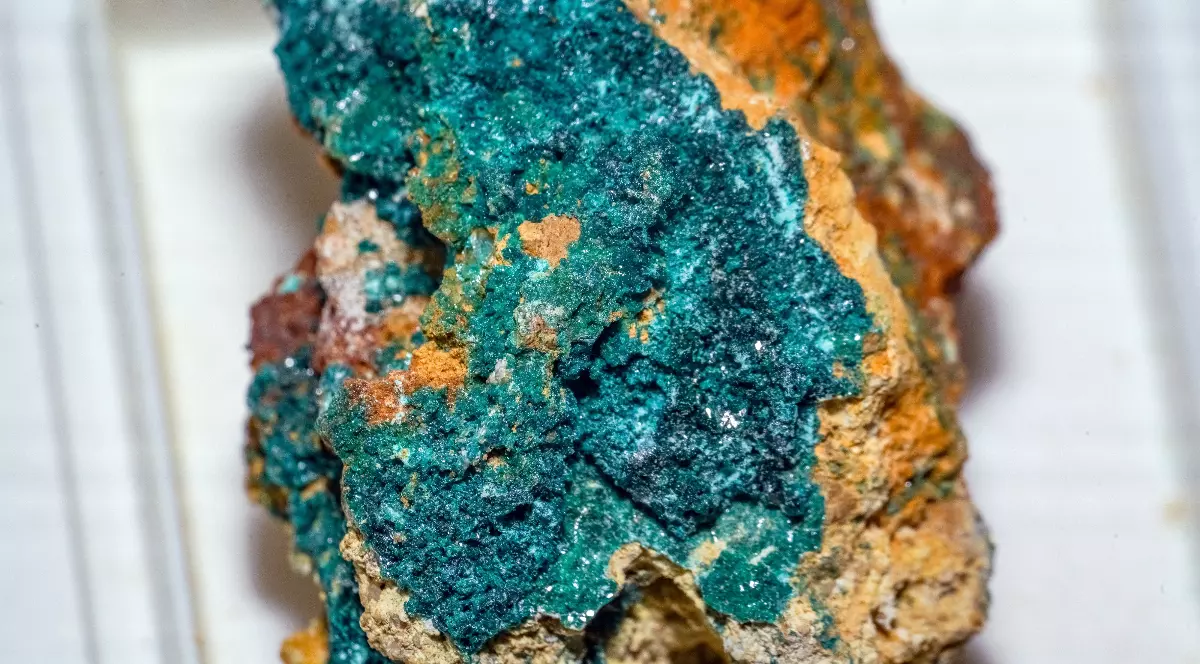 Imaginea prezintă un specimen de herbertsmithită, un mineral de culoare verde-albăstruie, expus pe o matrice de rocă maronie.
