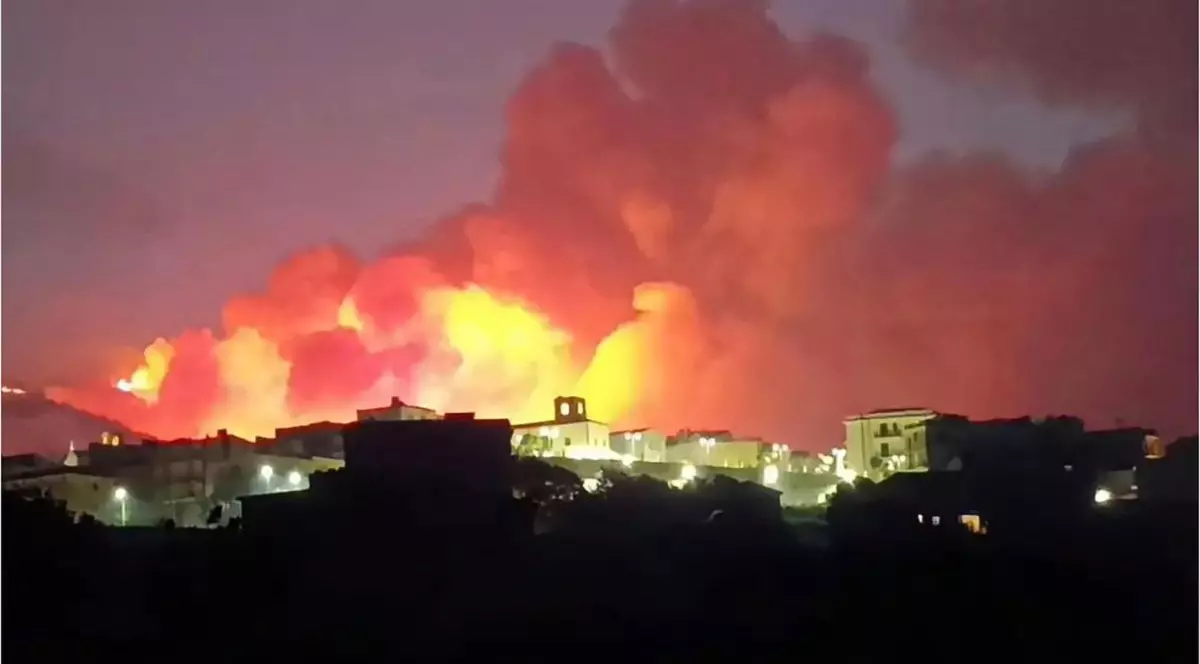 o așezare umană pe timp de noapte, profilată pe fundalul unui incendiu de proporții. În partea inferioară a imaginii se conturează siluetele unor clădiri de diferite dimensiuni, așezate pe un teren colinar, printre care se observă surse punctiforme de iluminat stradal. Imediat în spatele acestor construcții se ridică volume masive și dense de fum, care domină restul cadrului. Baza acestor formațiuni de fum este puternic luminată de flăcările intense ale focului care mistuie relieful din vecinătatea caselor, creând un contrast evident cu clădirile din prim-plan aflate în penumbră.