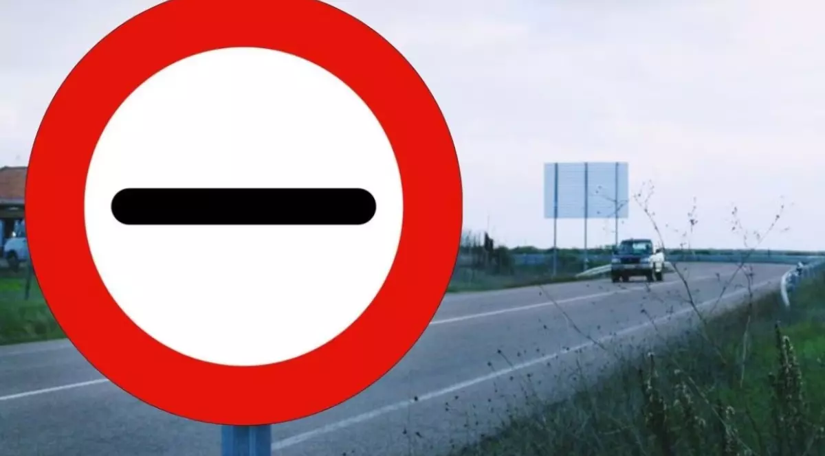 Imaginea prezintă în prim-plan un indicator rutier circular, alb cu margine roșie și o linie orizontală neagră în centru („Trecerea fără oprire interzisă”), amplasat pe marginea unui drum pe care se deplasează un vehicul.