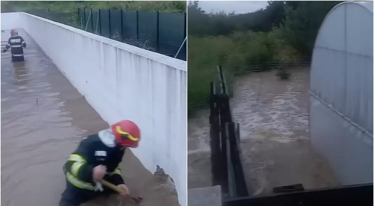 Imaginea surprinde intervenția pompierilor într-o zonă inundată, unde apa a acoperit curți și alei în urma ploilor abundente. În partea dreaptă se vede o gospodărie afectată de viitură, iar în stânga un pompier încearcă să elibereze scurgerea pentru evacuarea apei.