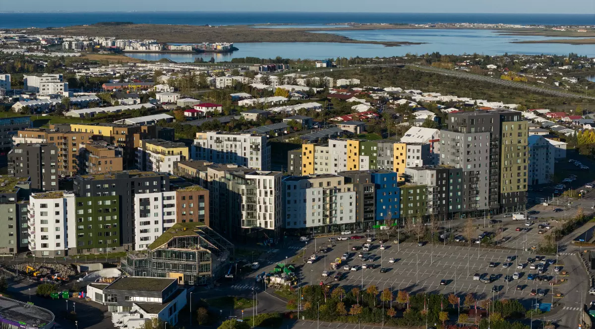 Imaginea surprinde o panoramă a unui cartier modern din Reykjavík, cu blocuri rezidențiale, clădiri de birouri și infrastructură urbană bine dezvoltată. În fundal se observă litoralul și întinderile de apă care înconjoară capitala Islandei, oferind un contrast între peisajul urban și cel natural.