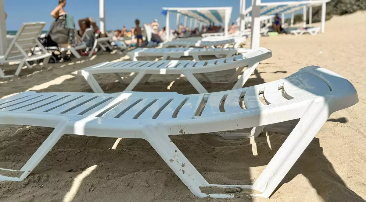 Mai multe șezlonguri din plastic alb, plajă în fundal, puse unele lângă altele pe nisip, în stânga se văd câțiva oameni în picioare și o apă în fundal