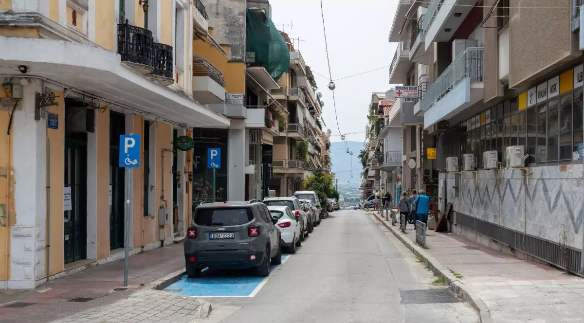 Stradă comercială în pantă, cu mașini parcate, locuri de parcare pentru persoane cu dizabilități, magazine și pietoni, în centrul orașului Lamia.