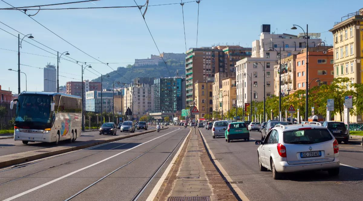 Imaginea prezintă un bulevard urban aglomerat, cu mașini și un autocar în trafic, mărginit de clădiri de locuințe și cu un deal pe fundal.