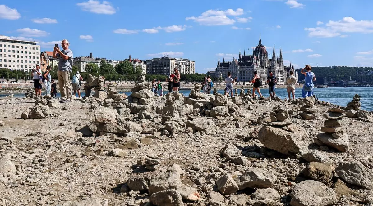 lbia secată a Dunării în Budapesta, oferind o perspectivă inedită asupra orașului. În prim-plan și în planul median se întinde o suprafață vastă și accidentată, acoperită cu numeroase pietre de dimensiuni variate, bolovani și resturi de rocă, indicând un nivel extrem de scăzut al apei. Pe această întindere expusă, vizitatorii au construit o multitudine de mici turnulețe din pietre suprapuse, creând un peisaj texturat. Mai multe persoane sunt surprinse plimbându-se direct pe albia uscată, unii stând în picioare sau fotografiind împrejurimile cu telefoanele mobile. Dincolo de această zonă pietroasă și de o porțiune mult îngustată a fluviului, pe malul opus, domină peisajul arhitectural clădirea monumentală a Parlamentului maghiar, o structură grandioasă caracterizată printr-o cupolă centrală proeminentă și zeci de turnuri ascuțite. În stânga acesteia se înșiruie alte clădiri urbane masive cu mai multe etaje, așezate de-a lungul falezei presărate cu rânduri de copaci.