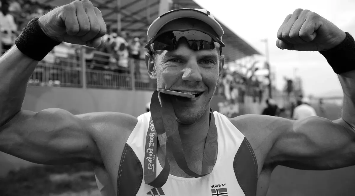 Imaginea îl surprinde pe un canotor pe podiumul de după o competiție, purtând o medalie la gât și ridicând brațele într-un gest de victorie.
