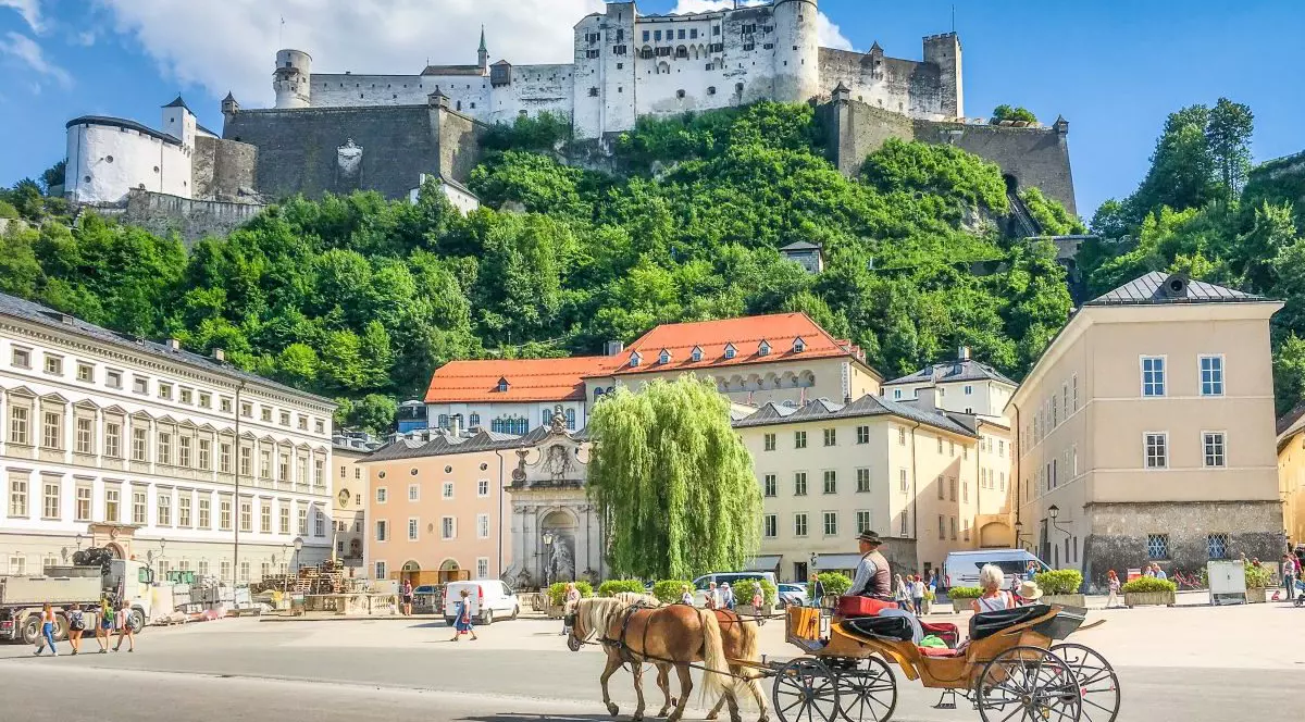 Vedere panoramică a orașului istoric Salzburg, cu trăsura tradițională Fiaker trasă de cai și faimoasa fortăreață Hohensalzburg pe un deal, într-o zi însorită cu cer albastru și nori de vară