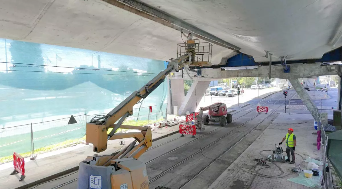 Imaginea surprinde lucrări de reabilitare desfășurate sub un pasaj rutier, unde mai mulți muncitori utilizează platforme ridicătoare și echipamente pentru intervenții la structura de beton. Zona este delimitată cu garduri și bariere de protecție, iar circulația pe liniile de tramvai și pe carosabil este restricționată pe durata lucrărilor.