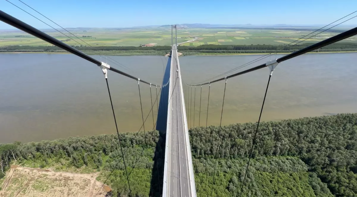 Imaginea surprinde un pod suspendat care traversează un fluviu lat, fotografiat de la mare înălțime, punând în evidență cablurile de susținere și perspectiva impresionantă asupra construcției. De o parte și de alta a apei se văd păduri și terenuri agricole întinse, într-un peisaj dominat de cer senin.