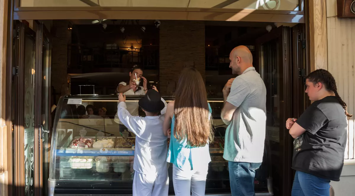 Imaginea surprinde mai mulți clienți așteptând la vitrina unei gelaterii, în timp ce un angajat le servește înghețată.