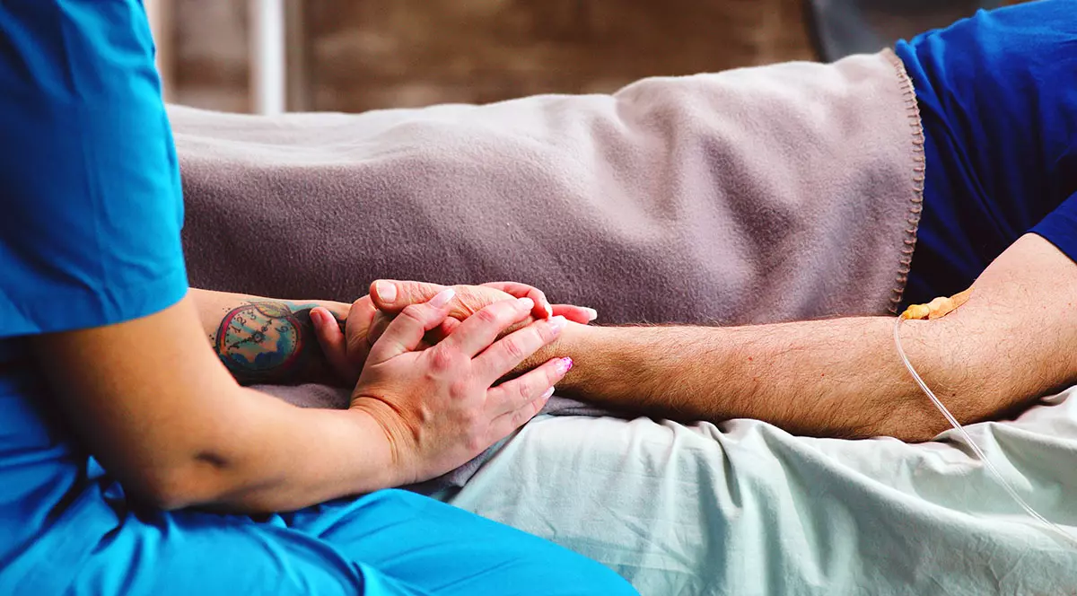 O asistentă ține de mână un bolnav întins pe patul de spital.
