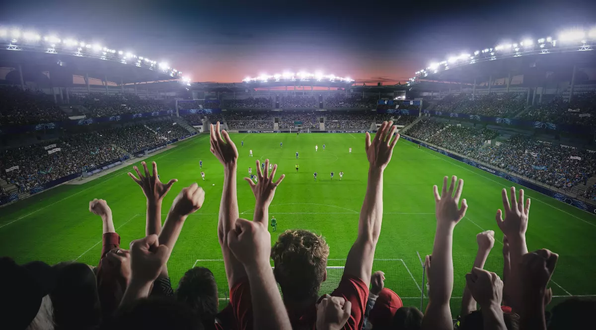 Imaginea surprinde un stadion de fotbal plin de spectatori, într-o atmosferă electrizantă, în timpul unui meci disputat seara sub lumina reflectoarelor. Suporterii din prim-plan își ridică mâinile și încurajează echipa, în timp ce jucătorii se află pe teren în plină desfășurare a partidei.