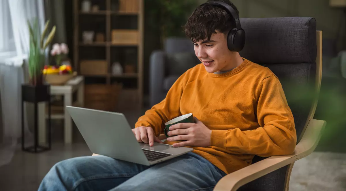 Un elev care stă pe un fotoliu, cu un laptop în brațe și citește ceva pe ecran. Poartă un pulover portocaliu și ține o cană de ceai în mână