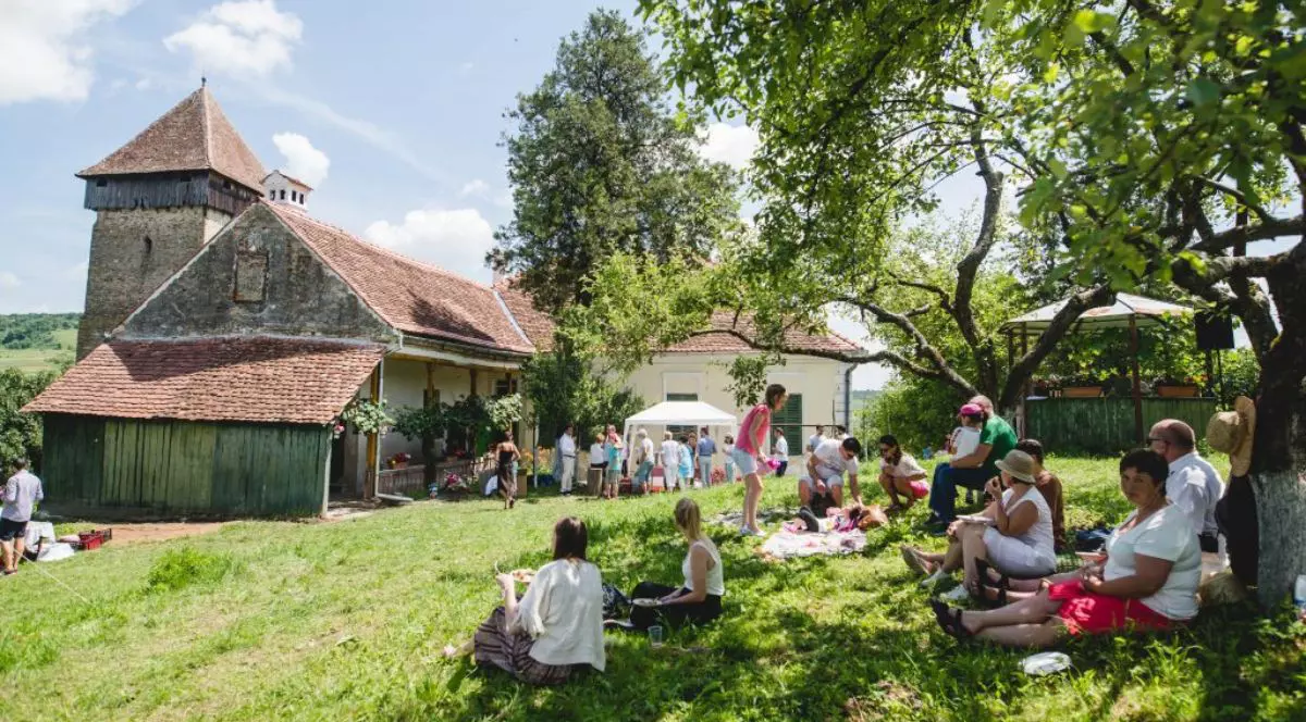 o scenă animată de relaxare în aer liber, desfășurată într-o curte spațioasă acoperită cu iarbă. În partea stângă se înalță o clădire veche, cu ziduri masive, acoperiș din țiglă și un turn robust, sugerând o arhitectură istorică de tip fortificat, având atașată o anexă cu pereți din scânduri. Partea dreaptă a cadrului este dominată de trunchiul și crengile bogate ale unui copac matur, care oferă o umbră generoasă. Numeroase persoane participă la această adunare; în prim-plan, în zona umbrită din dreapta, mai mulți oameni stau așezați direct pe iarbă sau pe pături, interacționând sau luând masa. În plan secund, mai aproape de clădiri și de un mic cort tip pavilion instalat în centrul curții, se observă un alt grup numeros de participanți care stau în picioare, socializând.