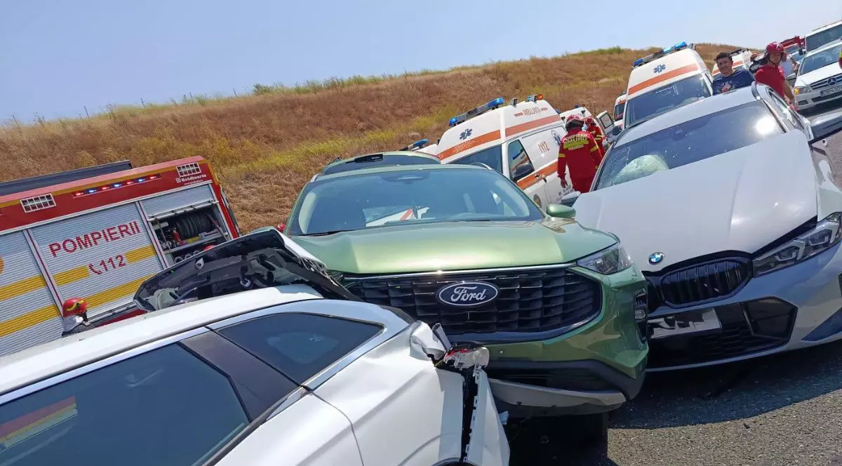 accident rutier în lanț pe o șosea mărginită de un taluz de pământ acoperit cu vegetație. În prim-plan este înfățișată o serie de autoturisme puternic avariate și încastrate unele în altele, având elemente ale caroseriei contorsionate, faruri sparte și bare de protecție smulse în urma coliziunilor. În spatele acestui carambol sunt staționate mai multe vehicule de urgență, printre care o autospecială de pompieri cu compartimente de echipament deschise și mai multe ambulanțe. În spațiul dintre mașinile avariate și autospeciale se află pompieri și paramedici în uniforme de intervenție, alături de martori, evaluând pagubele și desfășurând operațiuni de salvare.