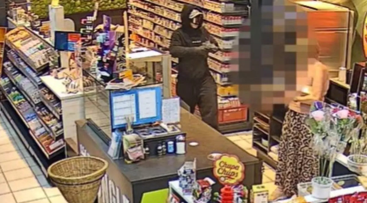 O imagine surprinsă de o cameră de supraveghere arată o persoană mascată și îmbrăcată în negru care comite un jaf armat într-un magazin, îndreptând o armă spre persoana de la casă.