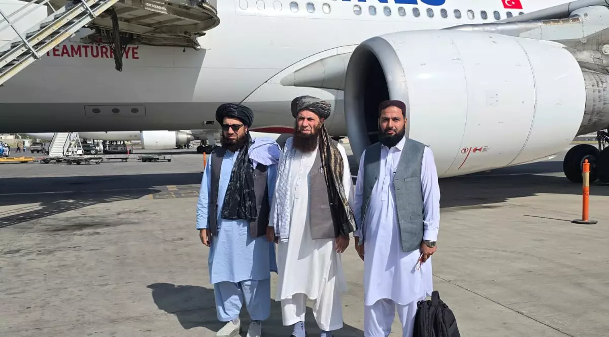 În imagine sunt trei bărbați cu barbă, îmbrăcați în haine tradiționale, care pozează pe pista unui aeroport. În spatele lor se află o aeronavă comercială de mari dimensiuni, parcată lângă scările de îmbarcare.