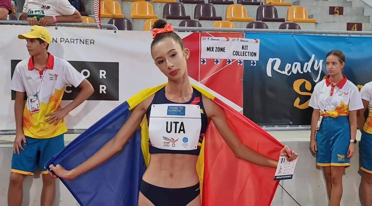 Alexandra Ștefania Uță, în echipament de atletism, cu steagul României pe spate și tribunele pe fundal