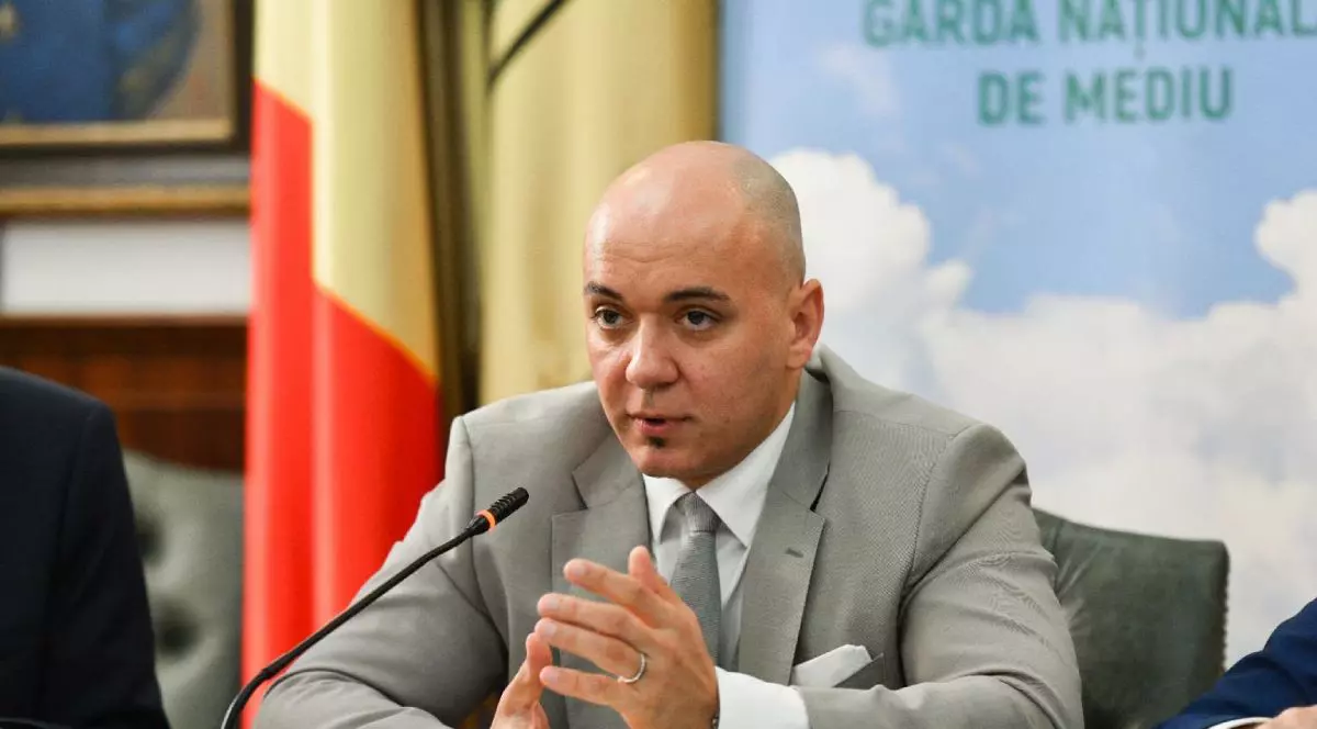Andrei Corlan, șeful Gărzii Naționale de Mediu, îmbrăcat cu un costum de culoare gri, cu o cămașă albă și cravată gri, cu steagul României pe fundal și un microfon în față, în timp ce stă la o masă, așezat pe un scaun