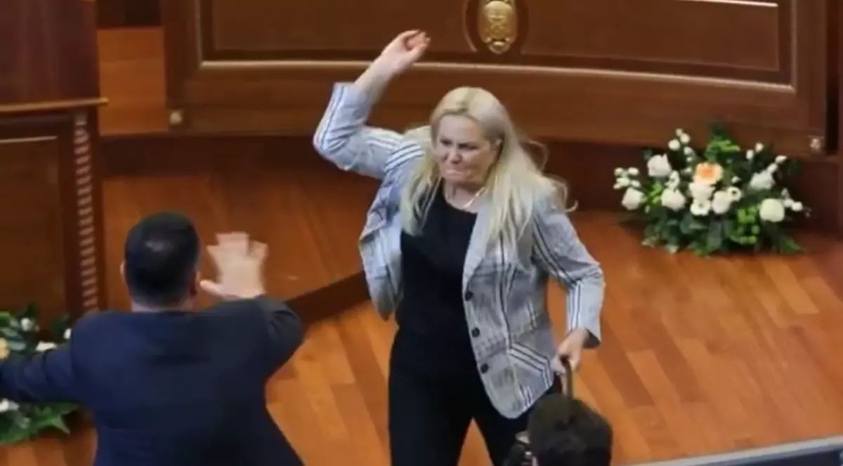 Imaginea prezintă o femeie blondă într-un sacou care ridică mâna pentru a arunca un obiect într-un bărbat dintr-o sală de parlament, în timp ce acesta încearcă să se apere.
