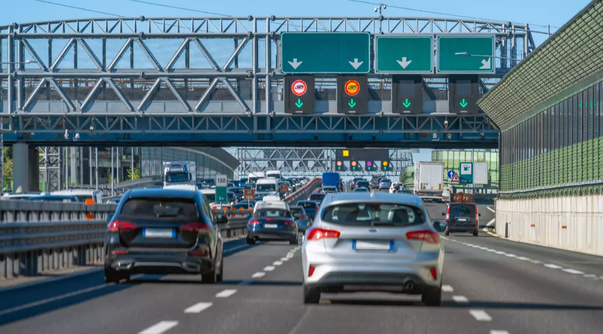 Imaginea prezintă un trafic aglomerat de autoturisme pe o autostradă, trecând pe sub o structură metalică masivă dotată cu panouri electronice și indicatoare rutiere.