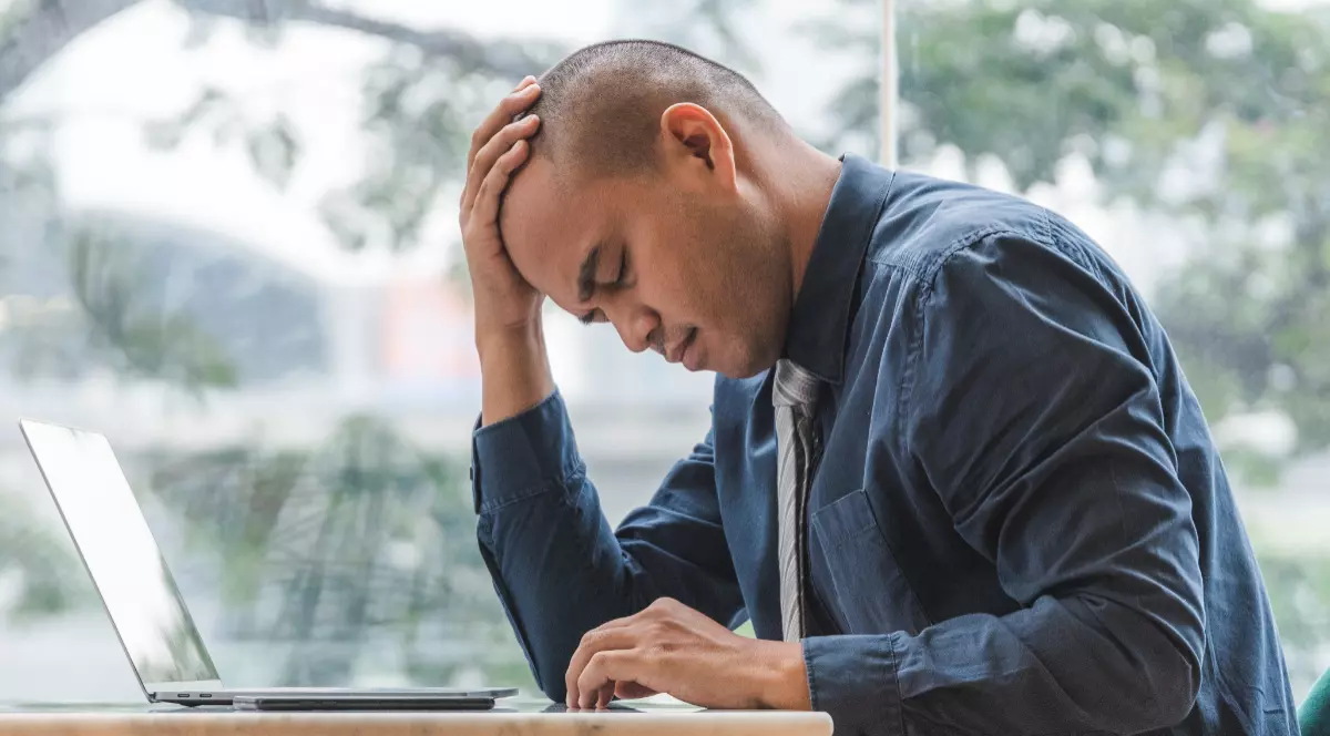 Un bărbat care stă la birou, scrie la laptop și se ține cu mâna de cap. Acesta este tuns scurt și poartă o cămașă gri