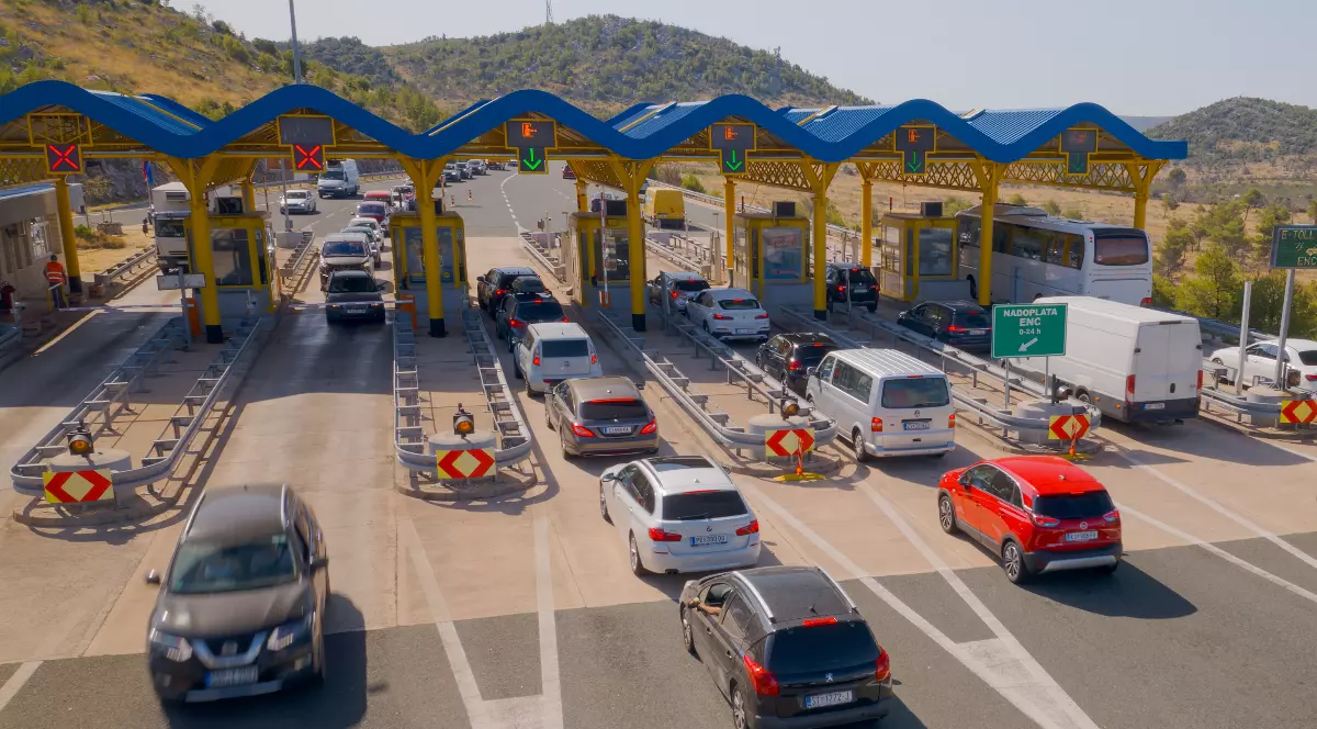 Imaginea prezintă o stație de taxare pe o autostradă, unde numeroase vehicule se află la coadă pentru a trece prin cabinele de plată.