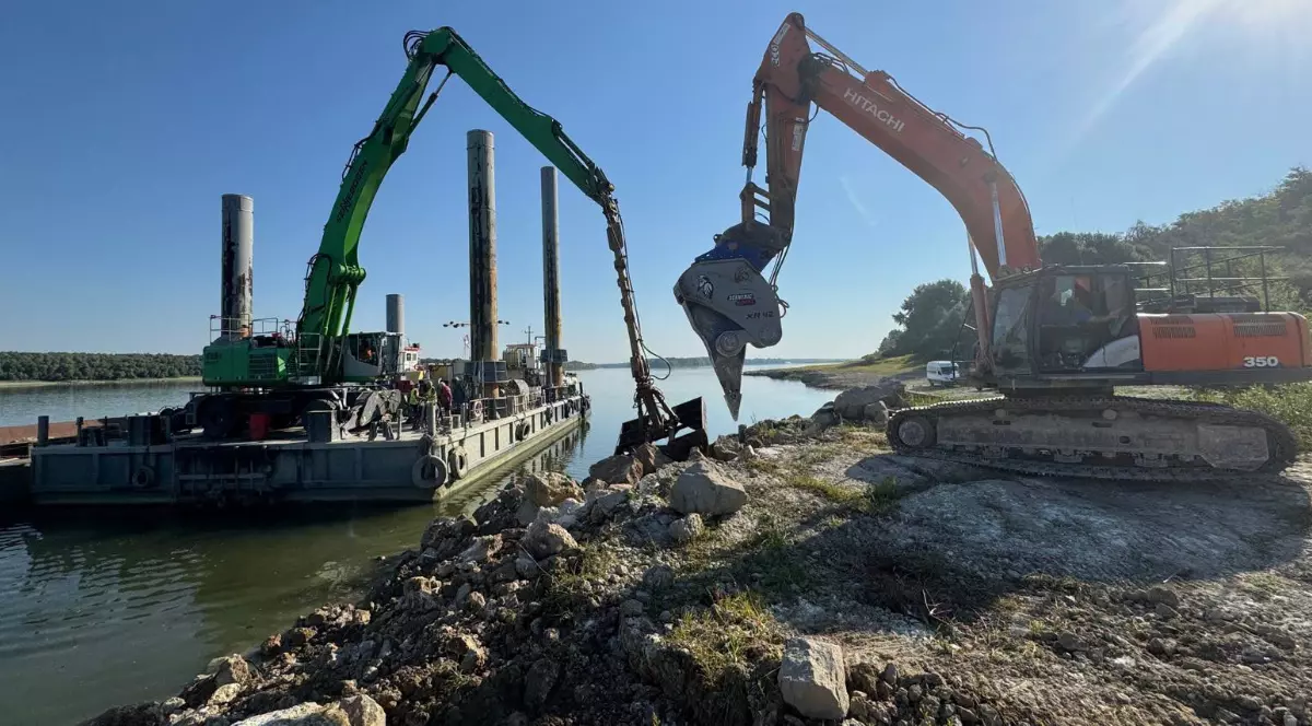 Un excavator care sapă și încarcă pietriș de pe malul Dunării pe o barjă
