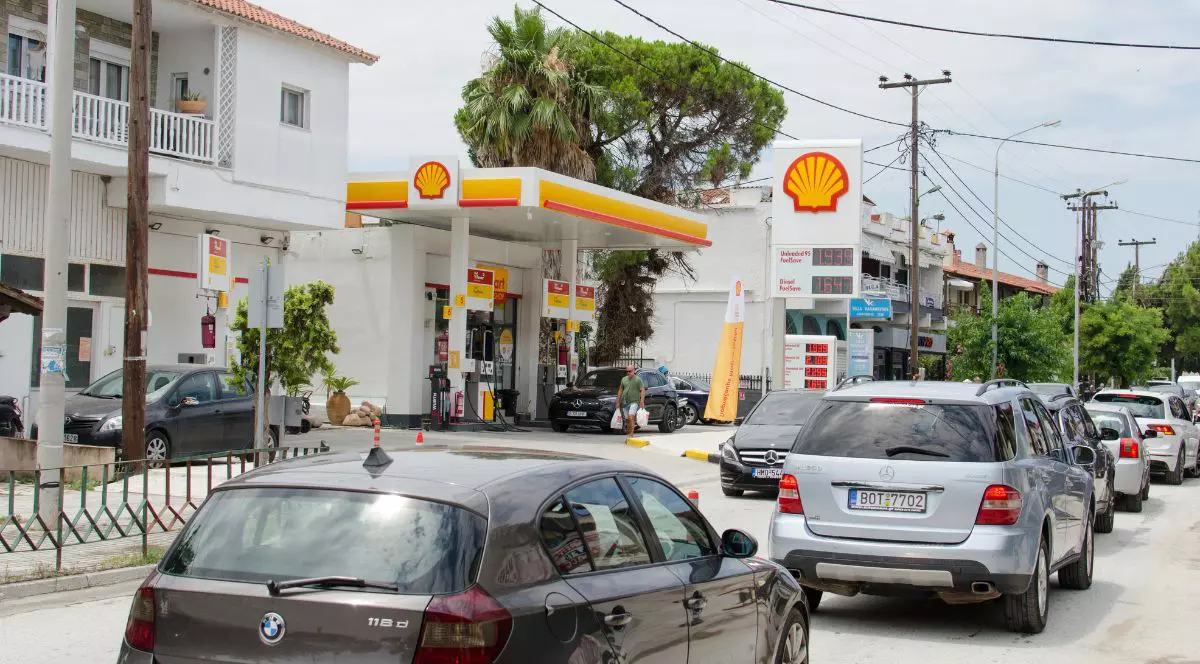 Imaginea surprinsă în Halkidiki, Grecia, prezintă o stradă intens circulată, mărginită de clădiri cu etaj și vegetație bogată, printre care se distinge trunchiul și coroana unui palmier. În prim-plan, carosabilul este ocupat de o coloană compactă de autoturisme oprite în trafic, ieșind în evidență o mașină cu formă rotunjită în partea stângă și un vehicul de teren în dreapta. Pe partea stângă a străzii, integrată în peisajul urban, se află o stație de alimentare cu carburanți. Aceasta este alcătuită dintr-un acoperiș plat susținut de stâlpi groși, sub care sunt amplasate pompele de alimentare, și un panou informativ foarte înalt pe care tronează o siglă în formă de scoică și lista de prețuri. În spațiul benzinăriei sunt parcate alte vehicule, iar un pieton este surprins traversând zona pompelor, în timp ce de-a lungul întregii străzi se înșiruie stâlpi de utilități interconectați prin cabluri aeriene.