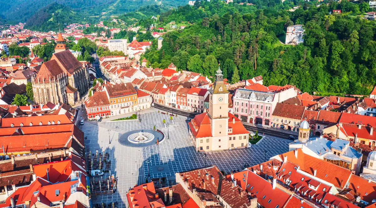 Imaginea surprinde o vedere aeriană a centrului istoric al Brașovului, cu Piața Sfatului, Casa Sfatului și Biserica Neagră în prim-plan. În fundal se întind dealurile împădurite care înconjoară orașul, oferind un contrast spectaculos cu acoperișurile roșiatice ale clădirilor istorice.