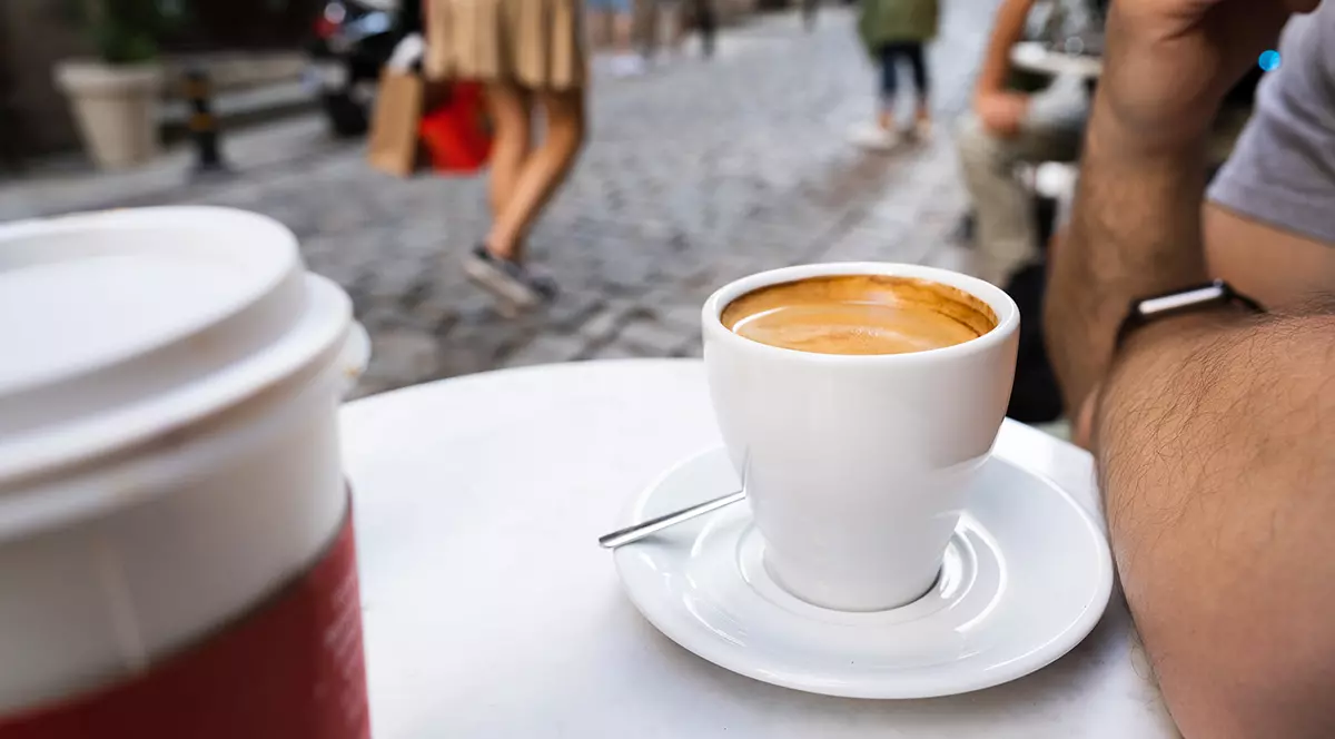 O cafea într-o ceașcă albă pe o măsuță iar în fundal se văd persoane care trec pe lângă.