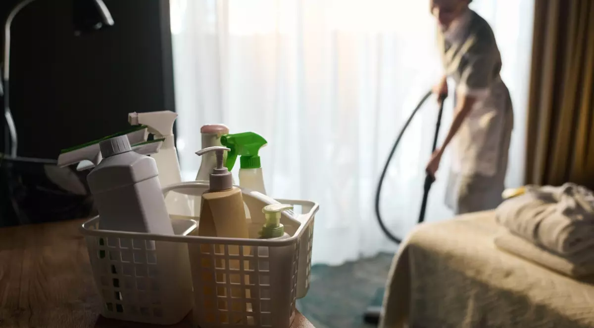 Imaginea prezintă în prim-plan un coș cu produse de curățenie, iar în fundal o cameristă în uniformă care aspiră podeaua unei camere de hotel.