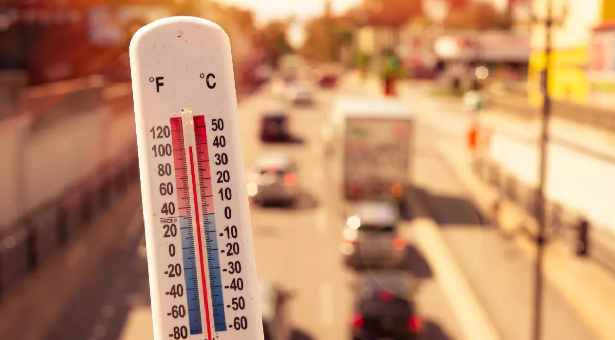Imaginea prezintă un termometru alb care indică o temperatură ridicată de aproape 40°C, pe fundalul estompat al traficului rutier dintr-un oraș în plină caniculă.