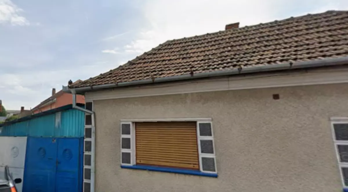În imagine se vede o casă cu un singur nivel, având fațada într-o nuanță deschisă și elemente decorative albastre în jurul ferestrelor și la baza pereților. Imobilul este prevăzut cu o poartă metalică albastră și un acoperiș din țiglă, iar în față se observă o alee asfaltată și o mașină parcată parțial în cadru.