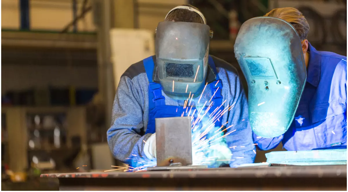 Doi muncitori echipați cu măști de protecție care rsudează într-un atelier industrial, în timp ce sar scântei luminoase.