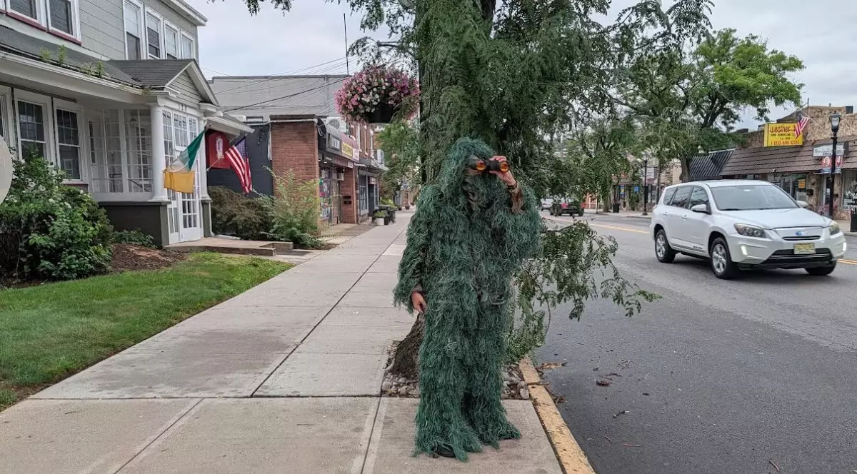 Imaginea surprinde o persoană îmbrăcată într-un costum de camuflaj verde, asemănător unui copac sau unui arbust, care privește printr-un binoclu pe o stradă dintr-o zonă urbană. Costumul acoperă aproape în întregime corpul și capul, iar în jur se observă copaci, magazine și autoturisme parcate.