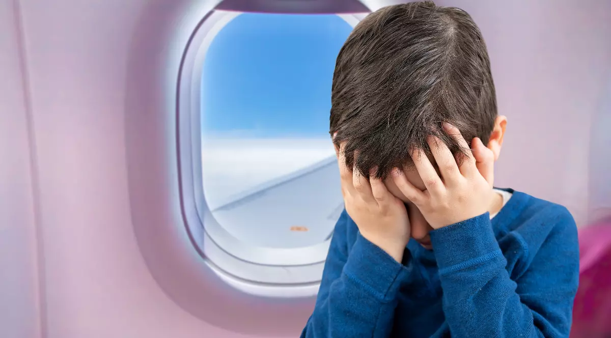 În imagine se vede un copil așezat într-un avion, lângă hublou, cu mâinile pe față și capul plecat. Expresia și postura sa sugerează că este supărat, copleșit sau stresat în timpul zborului.