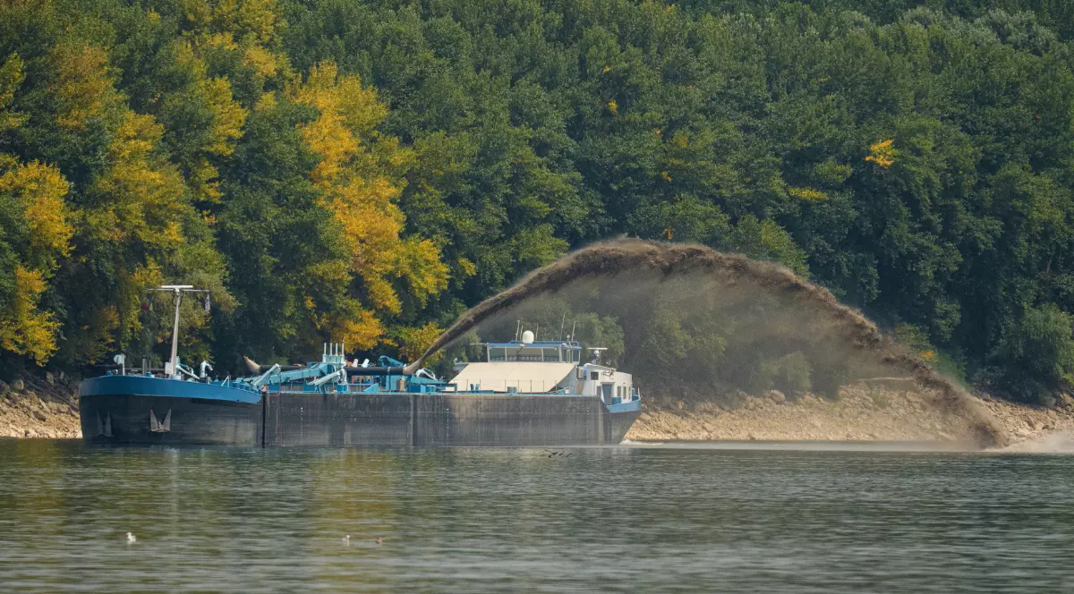 O navă de dragare desfășoară lucrări pe Dunăre, evacuând sedimente din albia fluviului pentru a îmbunătăți condițiile de navigație și debitul apei. În fundal se observă malul împădurit, în timp ce jetul de material extras este proiectat spre țărm.