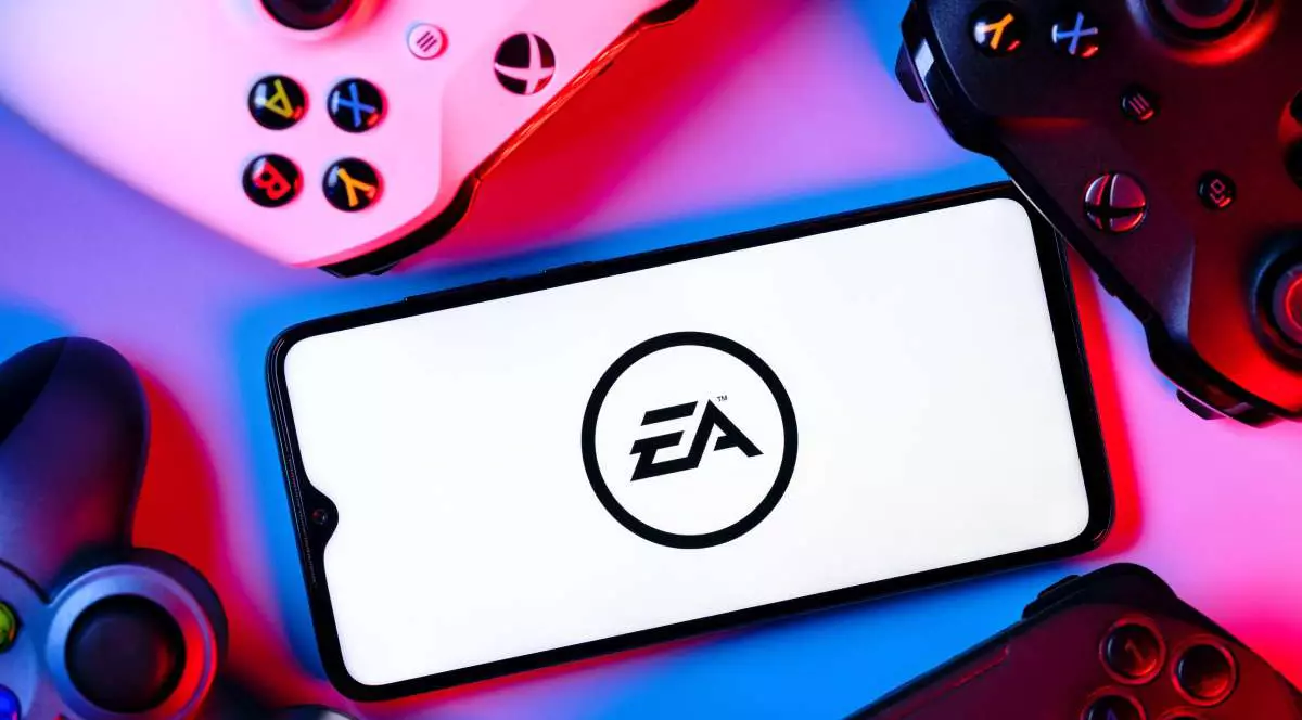 Logoul Electronic Arts, afișat pe ecranul unui smartphone așezat în mijlocul a mai multe controllere de PlayStation și Xbox.