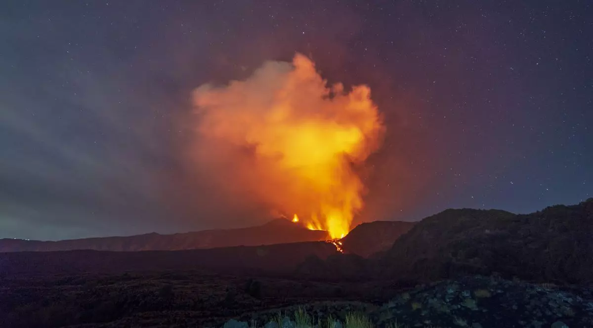 erupția vulcanului Etna din Italia, Sicilia, privit de la distanță. Din craterul situat pe creasta muntelui se înalță o coloană masivă și densă de fum, puternic luminată din interior de activitatea vulcanică. Pe versant se prelinge un râu de lavă incandescentă, iar în prim-plan se întinde un relief muntos accidentat, format din roci și vegetație măruntă.