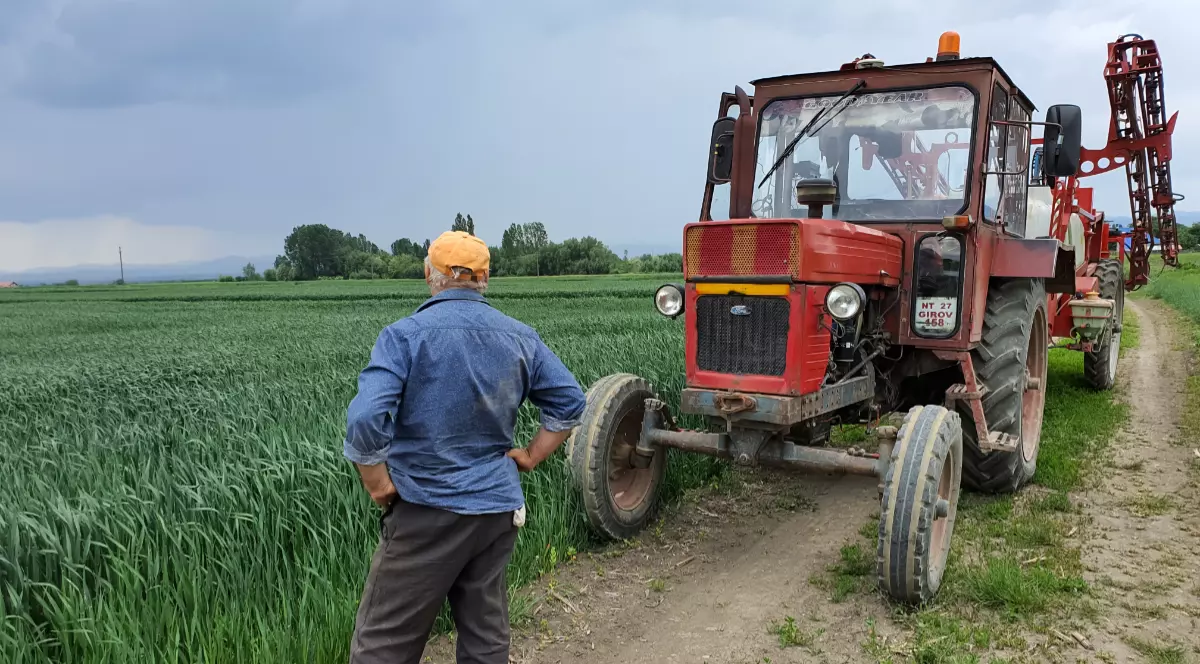 Un fermier care stă cu mâinile în șold și se uită la un lan de grâu, are lângă el un tractor