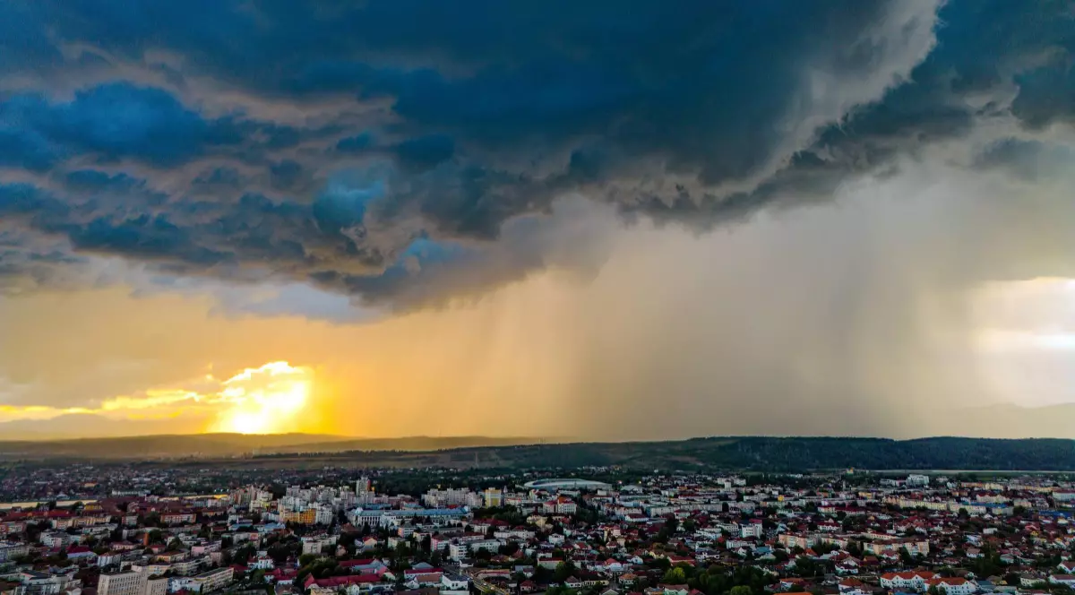 Imaginea prezintă o panoramă aeriană asupra unui oraș deasupra căruia se extind nori de furtună, în timp ce o perdea densă de ploaie este iluminată de lumina caldă a apusului la orizont.