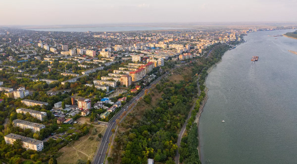 Imaginea surprinde o vedere aeriană spectaculoasă a orașului Galați, întins de-a lungul malului Dunării, cu numeroase blocuri, clădiri și spații verzi. În partea dreaptă se observă fluviul, pe care navighează o barjă, iar faleza și drumul de pe mal conturează peisajul urban.