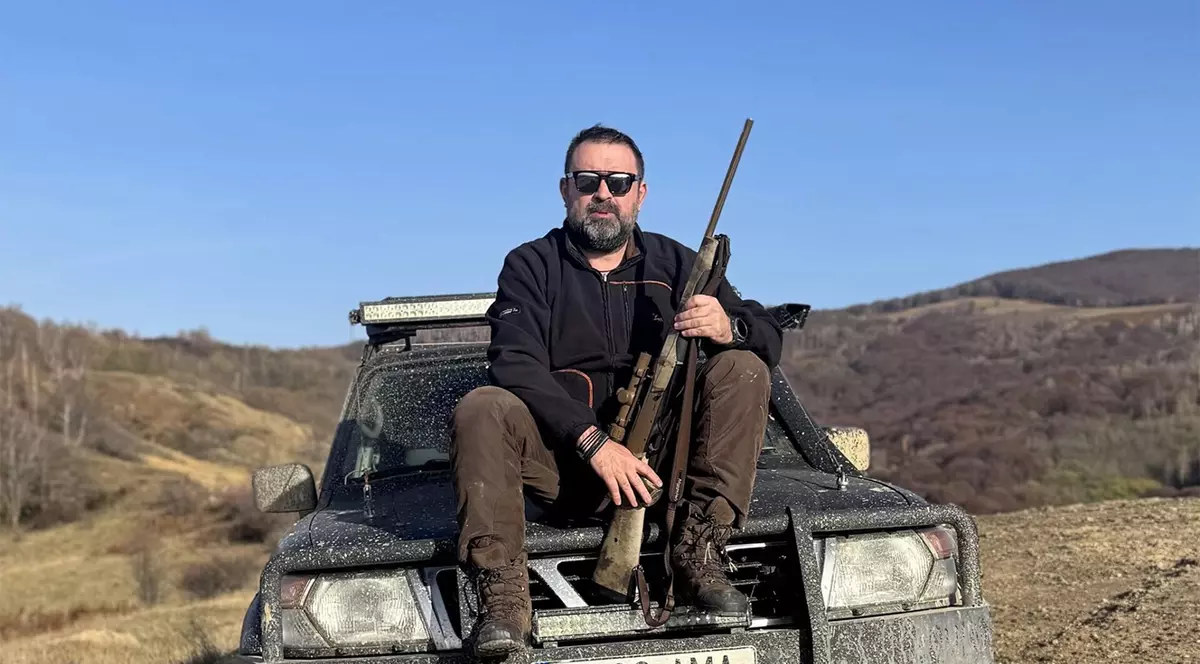 Un barbat cu o pusca in mana sta pe capota unei masini