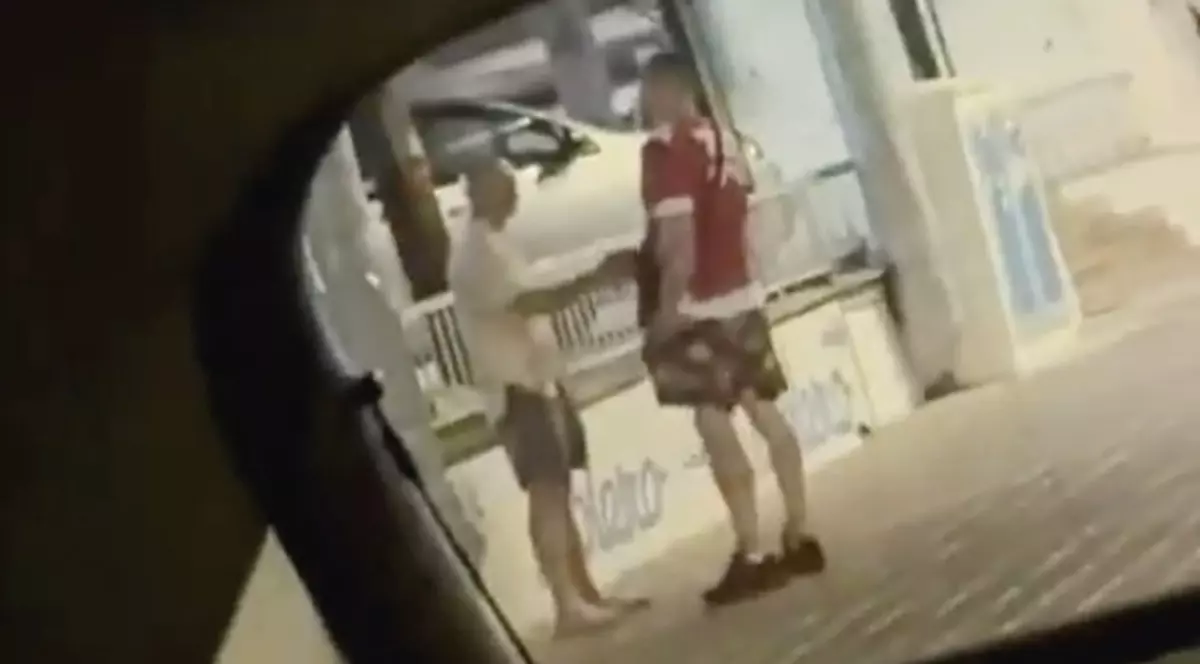 Imaginea prezintă doi bărbați care discută pe un trotuar pe timp de noapte, surprinși prin geamul sau oglinda unui vehicul.