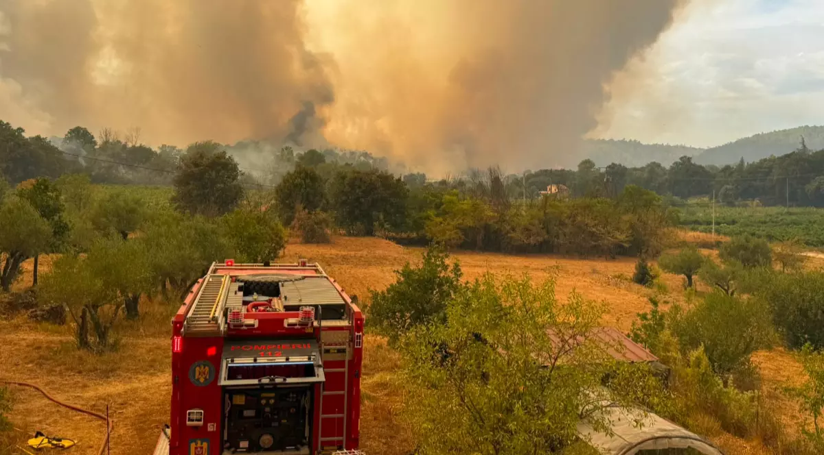 O mașină de pompieri pe un câmp din Grecia, în timpul unui incendiu de vegetație, în fundal se vede cum iese fum